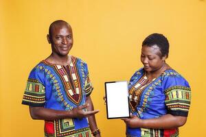 Black man and woman advertisers holding digital tablet with white screen, presenting new portable gadget. Smiling couple showcasing device with blank touchscreen mockup and looking at camera photo