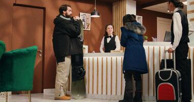Active people enter alpine resort lobby, approaching reception staff to see room reservation while carrying skiing equipment. Young couple at mountain hotel preparing for winter sport activity. photo