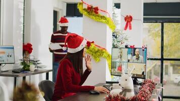 Asian worker in online videocall with colleagues during Christmas season in festive decorated office. Employee using computer webcam for remote teleconference with team in busy xmas ornate workplace photo