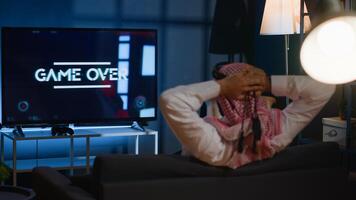 Man playing videogames on TV after long day at work angry after receiging game over screen. Muslim gamer upset after losing at science fiction shooter competition on gaming console photo