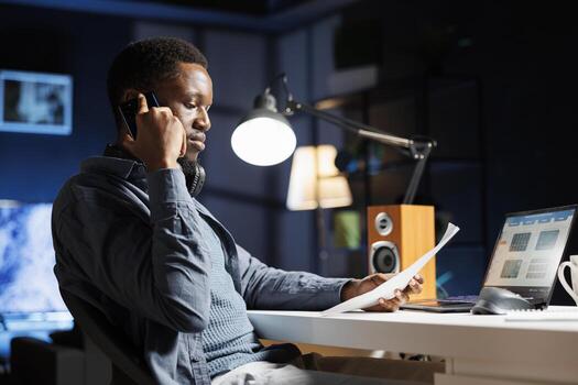 Working from home to expand enterprise on global stage, african american man calls creditors to acquire more money for capital expenditure. At night, staff member talks on mobile device. photo