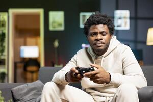 Man in rgb lit living room playing games on couch, enjoying day off from work. Gamer battling enemies in internet multiplayer shooter on gaming console, talking with teammates through headphones photo
