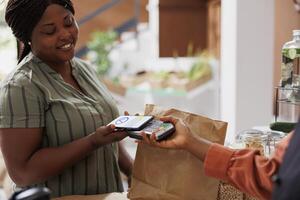 Cheerful black woman buying bio food with mobile payment at retail store checkout counter using credit card on her phone, while smiling and being served by a smiling vendor photo