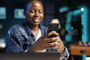 Smiling black woman using smartphone for texting while seated at desk with laptop. Close up of african american lady, working at home, clutching mobile device, browsing through social media on break. photo