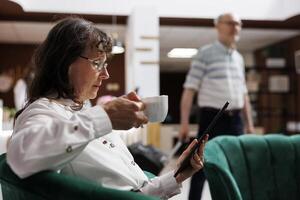 Side-view of eldery lady with coffee, browsing webpage on tablet for holiday activities. In hotel reception, senior woman in close up shot, holding a cup while surfing the net on her digital device. photo