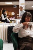 Professional hotel receptionist assists old man with check-in, verifying identity and retrieving reservation details. Concierge holds and checks personal document of male tourist. Focus on background. photo