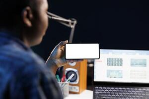 Close-up of freelancer working from home, using a laptop and smartphone displaying blank copyspace mockup template. Black woman looking at isolated white screen on mobile device. photo