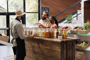 Male vendor offering advice to clients who are shopping for sustainable merchandise at eco friendly store. Owner of local supermarket describing food items in jars to the young multiracial couple. photo