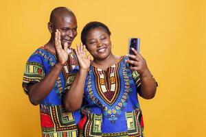 Cheerful black couple waving with hands while holding smartphone and speaking in online meeting together. Smiling man and woman pair saying hello while chatting in mobile phone videocall photo