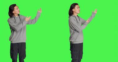 Asian guy doing advertisement on camera, pointing at something next to him for new marketing ad. Young adult smiling and working on promotional slogan, standing over greenscreen backdrop. photo