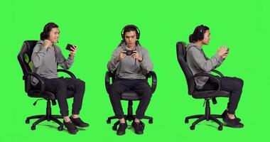 Asian guy sitting in chair over greenscreen, performing games on mobile device. Young man utilizing phone to engage in smartphone games, web roleplaying contest with players. photo