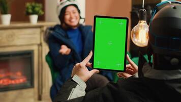 Person has tablet with greenscreen and talks to young man equipped in skiing attire, showing isolated copyspace with chromakey display on device. People in lounge area at alpine resort. photo
