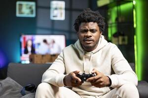 Bored person in living room plays online videogame, spamming attack buttons on gamepad. Gamer in RGB lit home participating in internet competition, sitting on couch and playing on gaming console photo