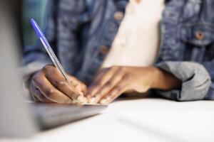 African American person works from home, using her laptop and taking notes on a notepad at a modern desk in her apartment. Close up of black person writing on a notebook, studying and researching. photo