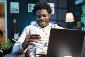 African american man laughing while adding payment method on website while in dimly lit living room. Joyous person delighted to do online shopping, looking at credit card in hand photo