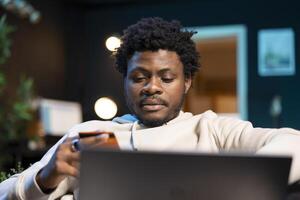 Carefree man preparing to do online shopping, carefully typing credit card information on laptop. African american person adding payment method on website while in neon lit home photo