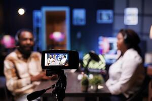 Detailed view of professional camera used to film live broadcast between show presenter and man in home studio. Closeup of production equipment with host and guest chatting in blurry backdrop. photo