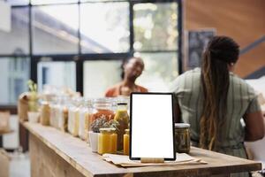 Tablet with isolated white screen vertically positioned near glass containers having organic products. While client and vendor converse, digital device displays blank mockup for bio food store ads. photo