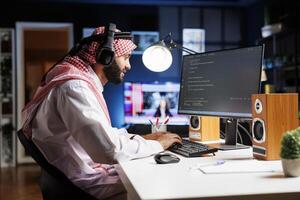 Middle Eastern software programmer typing code on a laptop showcasing the essence of software programming and development. Arab man with wireless headphones engrossed in running algorithms. photo