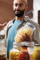 Close-up shot of middle eastern man grasping a container filled with homemade pasta in eco friendly store. Detailed image of male customer holding glass jar while browsing for other organic products. photo