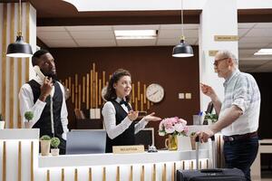 Friendly concierge checking senior man in at the main desk of a luxury hotel. While luggage is close in the lounge area, a pleasant grin from a female employee helps an elderly male tour guide. photo