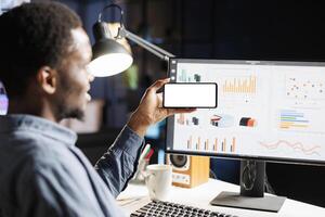 Remote worker uses smartphone with white display at desk, examining revenue target on web statistics. Young person holds mobile phone showing isolated empty screen, remote tasks. photo