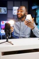 Smartphone on holder capturing footage of BIPOC man reviewing smart light bulb. Viral content creator talking with fans, filming IoT home technology gear with high quality phone camera photo
