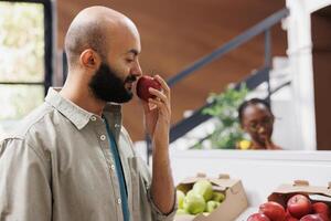 Middle eastern customer is seen smelling a red apple at local store. Arab man grasping and admiring the freshly harvested organic fruit at an environmentally friendly supermarket. photo