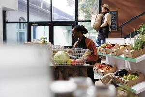 African American woman browses around a table where several reusable containers are filled with organic food products. Multicultural shopkeepers moving around local store, arranging sustainable items. photo