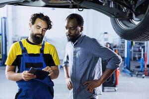 Seasoned mechanic in garage using laptop to follow checklist while doing maintenance on car, talking with client. Employee in auto repair shop does checkup on vehicle tires next to customer photo