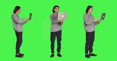 Modern man navigating webpage on laptop standing over full body greenscreen backdrop, browsing online website using wireless pc. Young guy checking social media page working on business. photo