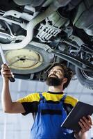Licensed mechanic working on suspended car in garage, checking wheels during routine maintenance. Qualified auto repair shop employee walking underneath vehicle, inspecting it using work light photo