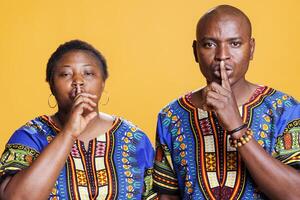 African american man and woman holding forefinger on lips and asking to keep secret portrait. Couple making shh gesture, showing silence signal and looking at camera with serious expression photo
