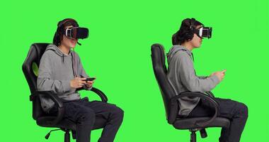 Gamer uses virtual reality for gameplay with mobile phone app, sitting on gaming chair in isolated greenscreen studio. Asian person playing multiplayer action games with vr. photo