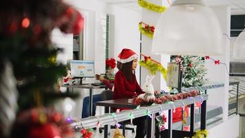 Joyful asian employee working on christmas season in festive decorated office. Smiling staff member solving tasks at desk in diverse xmas ornate workplace next to BIPOC colleagues photo
