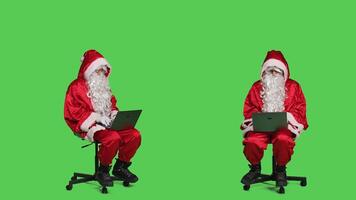 Cool saint nick using laptop on chair, sitting against full body greenscreen backdrop in studio. Father christmas working with modern portable pc, browsing online website in red costume. photo