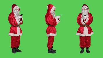 Father christmas using smartphone on camera, browse onine webpage on greenscreen backdrop. Modern santa character checks messages and internet sites, wears festive red costume. photo