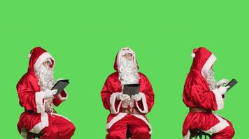 Saint nick using tablet in studio sitting on chair against greenscreen background, working on modern device to create presents list. Modern santa claus browsing website on gadget. photo