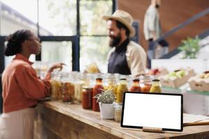 Tablet with a blank chromakey mockup template horizontally positioned near glass jars. On the counter is a smart device displaying an isolated white screen while shopkeeper converses with customer. photo