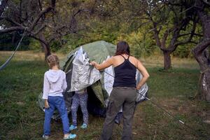mother and little daughters are setting up a tourist tent in forest, camping with children photo