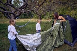 mother and little daughters are setting up a tourist tent in forest, camping with children photo