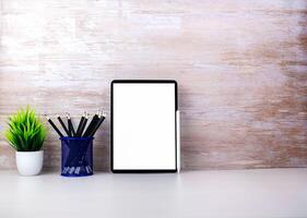 Tablet with blank screen placed on white table and leaning against wooden surface wall with office equipment. Copy space for your text. Workspace and copy space. Office desk with office supplies photo