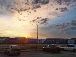 The horizon glows with orange and yellow hues as the sun sets behind clouds, casting a warm light on nearby parked cars and the cityscape in Puncak Alam Malaysia. photo