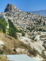 The landscape showcases Cappadocia's iconic rock formations and rolling hills, with lush greenery and distant historical sites visible under bright sky. photo