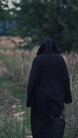 Backside view of witch on the field. Black death goes slowly away along the path on nature background. Vertical video