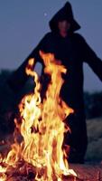 Fire and black figure at darkness. Spooky death in cloak with hood standing against the bright flame and doing some ritual outdoors. Halloween. Vertical video