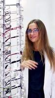 Pretty customer in eyewear store. Lovely girl in eyeglasses standing near the stand with new spectacles and looking at camera. Eyesight concept. Vertical video