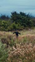 Scary figure of death in the field. Ghost in black cloak standing alone with outstretched arms by sides on nature background. Vertical video