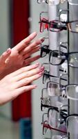 Customer choosing new glasses in the store of eyewear. Young woman turning stand with fashionable spectacles at optics. Eyeglasses choice. Vertical video