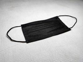 A black disposable face mask lies flat on a cream white surface. The image captures the simplicity and functionality of the face mask, showcasing its essential role in public health and safety. photo